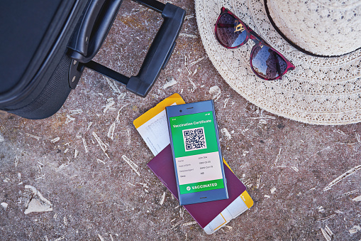 Smartphone displaying valid digital vaccination certificate for COVID-19, suitcase, summer hat, passport and boarding pass. Vaccination and health passport, international travel health control