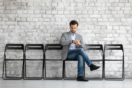 Worried job seeker waiting for interview far too long, sitting on chair alone in row, consulting watch. Male professional candidate expecting meeting with late employer or recruiter, checking time
