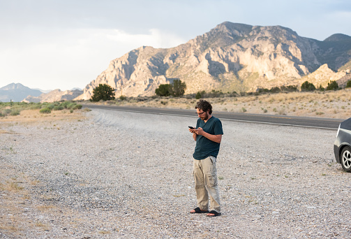 On a summer day a man driving cross country stops for a break to look at his mobile phone for cell phone reception in the rural desert of Nevada, USA.