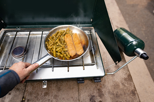 This is a photograph from a personal perspective of a woman’s hands holding onto a small frying pan as a breakfast of vegan sausage and green beans is being cooked outdoors on a camping stove in Mount Shasta, California.