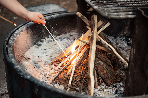 This is a photograph of a small fire being lit by an unrecognizable boy’s hand outdoors in a fire pit in summer at a Mount Shasta campsite in California, USA.