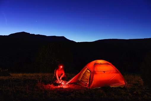 Hiker Camper Backpacker at Dusk with Glowing Orange Tent - Scenic mountain views during blue hour twilight of rugged backcountry setting.
