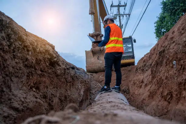 Engineer wear safety uniform examining excavation concrete Drainage Pipe and manhole water system underground at construction site.