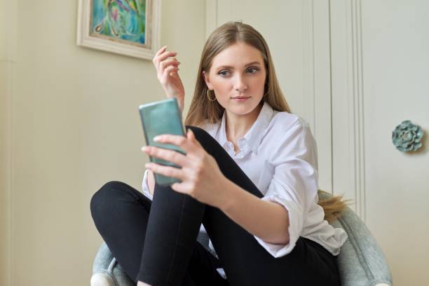 istock Young blonde sitting at home in chair, using video communication on smartphone 1318577410