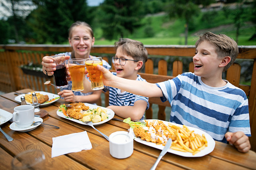 Teenage girl and her younger brothers are having lunch in outdoors restaurant. The kids are drinking juices eating chicken cutlets with French fries.\nNikon D850