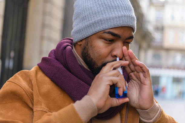 Young African-American Man with Protective Face Mask is Having Sinusitis Problems Outside in Nature. A Man of African-American Ethnicity is Using Nasal Spray Due to the Problems with Nose and Breathing in the Public Park During a Pandemic Time. nasal spray stock pictures, royalty-free photos & images