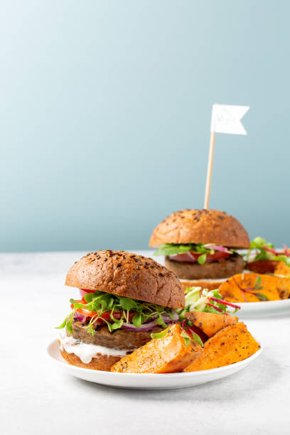 hamburguesa a base de plantas sin carne servida con cuñas de batata, ensalada de mezcla verde y salsa blanca sobre mesa gris cerca del fondo azul. concepto de comida vegana o vegetariana saludable. - sweet potato french fries yam baked fotografías e imágenes de stock