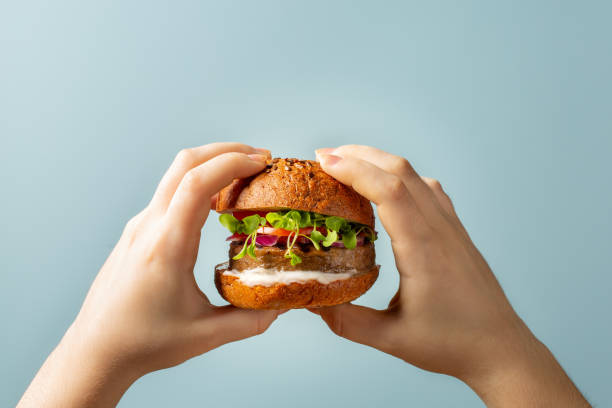 istock Hands holding burger. Hamburger with meat free plant based cutlet, tomatoes, onions, white sauce and microgreens on blue background. Healthy vegan or vegetarian food concept. 1318594064