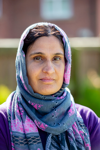 Close-up of a confident mid-adult woman wearing a headscarf smiling at the camera. The sun is shining. This is a British Muslim woman standing on her own.