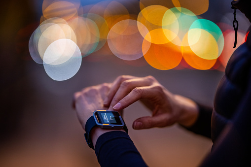 Close-up of female runner looking at her smart watch while taking a break from sports training. Woman monitoring fitness process on smartwatch in the city at night.