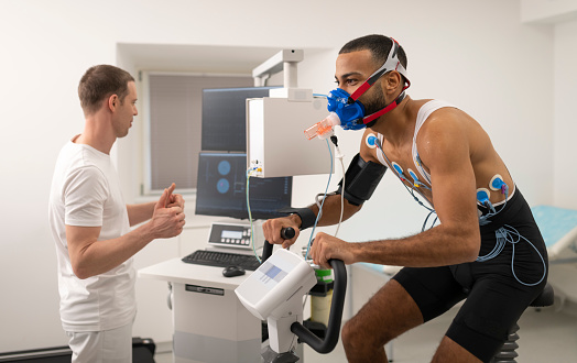 Male doctor observing the progress of a cardiopulmonary stress test taken by the male athlete riding a bicycle ergometer.