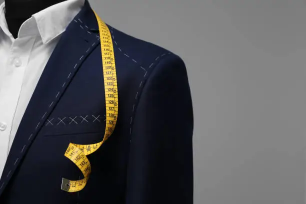 Photo of Semi-ready jacket with tailor's measuring tape on mannequin against grey background, closeup. Space for text