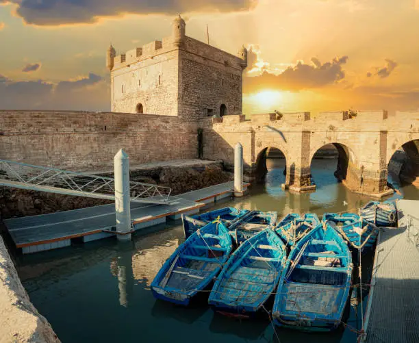 Historical fortress tower and walls and traditional blue fishing boats against the beautiful sunset in Essaouira in Morocco