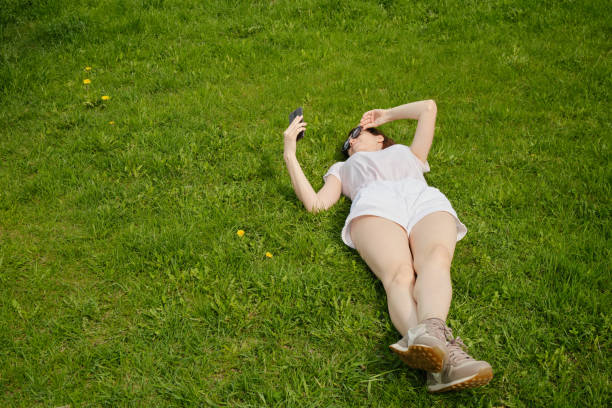 девушка в очках лежит на зеленой траве и держит смартфон - vacations photographing brown hair 30s стоковые фото и изображения