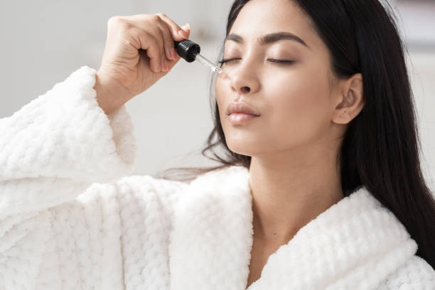 Young asian woman applying serum on facial skin Skincare and beauty concept. Portrait shot of young asian woman applying serum or essential oil on facial skin. Model in white bathrobe moisturizing derma with vitamin E, collagen and hyaluronic acid face serum stock pictures, royalty-free photos & images