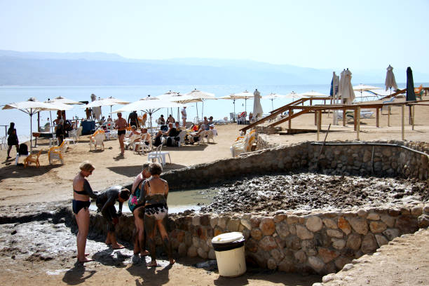 Horizontal photo of people coating themselves with mud from a mud pit next to a swimming area at the Dead Sea. The mud is prized for having rich minerals and healing properties for the skin.