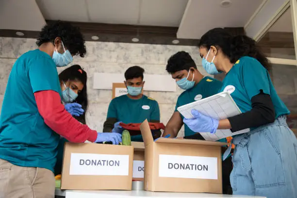 Photo of Low angle view, group of volunteers busy working by arranging vegetables and clothes on donation boxes and noting down during coronavirus covid-19 pandemic lockdown for needy people.