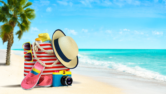Summertime vacations background. Beach accessories - beach bag, straw hat, flip flops and sunglasses on sandy beach with palm tree and azure sea on background