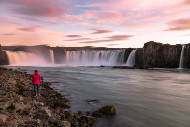 mulher caminhante em pé ao lado de um rio de fluxo rápido admirando uma cachoeira majestosa calorosamente iluminada pelo sol da meia-noite - sol da meia noite - fotografias e filmes do acervo