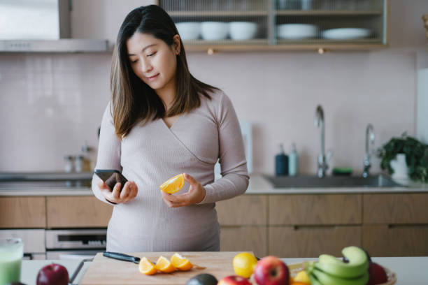 jonge aziatische zwangere vrouw die voedingsfeiten en calorieëninname van geassorteerd fruit met mobiele app op smartphone in keuken thuis controleert. gezonde voeding. goed eten. gezonde zwangerschap die levensstijl eet - pregnant count stockfoto's en -beelden