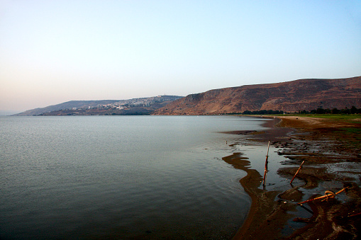 Horizontal landscape photo of the calm Sea of Galilee or Lake Tiberius, with the beach foreshore and distant hills under a cloudless pale blue sky in Spring