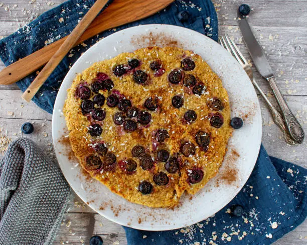 Brown rice pancake with fresh blueberries topped with grated coconuts and cinnamon served on a white plate on light wooden background from above