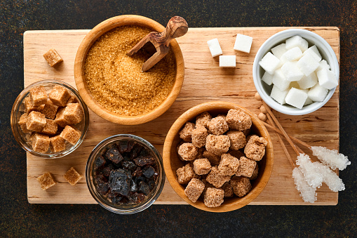 White sugar, cane sugar cubes, caramel in bamboo bowl on dark brown table concrete background. Assorted different types of sugar. Top view or flat lay.