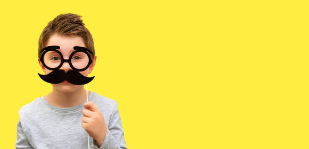 funny happy boy with black hipster mustache and glasses, child pretends to be dad, professor or nerd on yellow banner background, fathers day, mens health, smart kids, joke, emotion, copy space - costume mustache child disguise imagens e fotografias de stock