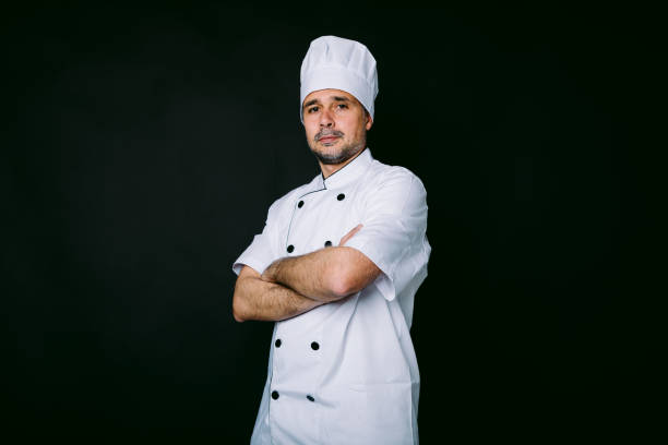 istock Chef cook wearing jacket and kitchen hat, poses with crossed arms on black background 1318652693
