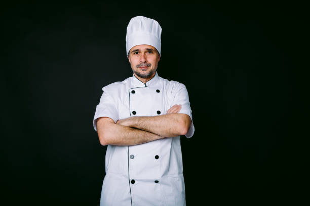 istock Chef cook wearing jacket and kitchen hat, poses with crossed arms on black background 1318652695
