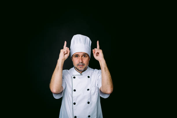 istock Chef cook wearing jacket and kitchen hat pointing up on black background 1318653461