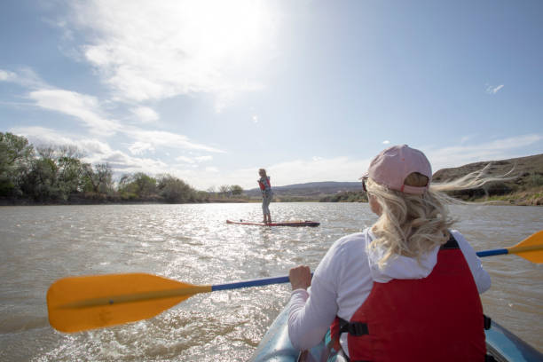 POV of woman kayaking down river along with paddle boarder She is wearing a baseball cap and life preserver fruita colorado stock pictures, royalty-free photos & images