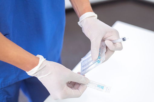 Shot of a nurses hands wearing white surgical gloves removing a needle from packaging. The nurse is prepping to inject a patient with the COVID-19 vaccine.