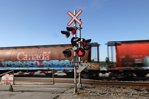 Maple Ridge, Canada - May 16, 2021: A freight train travels past flashing red lights at a level crossing at the foot of 225th Street in Maple Ridge. The track stretches alongside the Fraser River in Metro Vancouver.