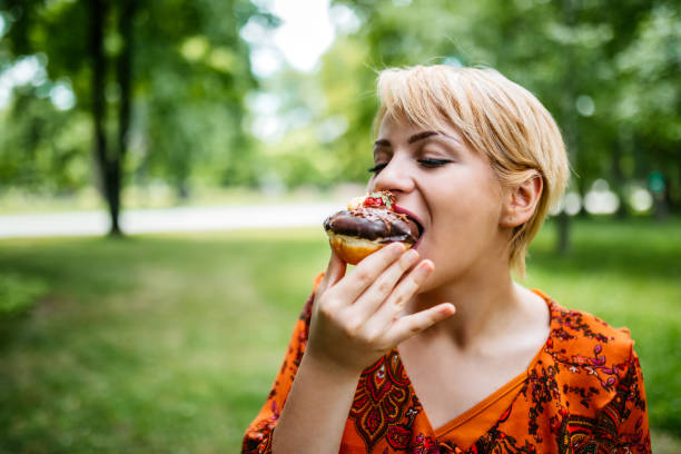 Young Caucasian cute woman enjoying eating donut in public park.