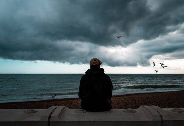 Lonely man looking at the sea and storm clouds Rear view color image depicting a young man sitting alone at the beach, looking at the dark sea and storm clouds. Moody image emphasising loneliness and isolation. Seagulls are circling and swooping in the distance. gloomy mood stock pictures, royalty-free photos & images