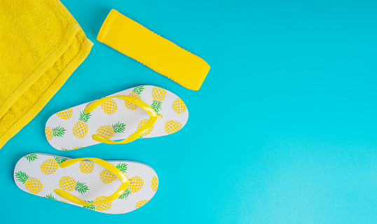White flip-flops thongs with pineapple decoration, bright yellow tube of sunscreen lotion and beach towel on vivid blue backdrop. Summer season vacation concept. Image with copy space, horizontal