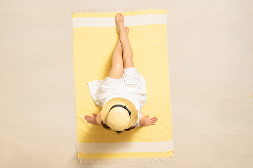 Alone woman in straw hat and dress sitting on yellow beach towel. Female relaxation on the sand of the beach at summer vacation. Top, aerial view
