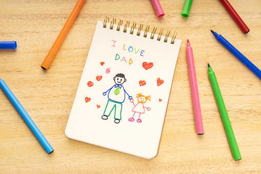 Fathers day concept. Notebook with drawing of a father with a girl and marker pens on a wooden table