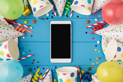 Top view photo of birthday composition party accessories and smartphone with copyspace in the middle on isolated blue wooden table background