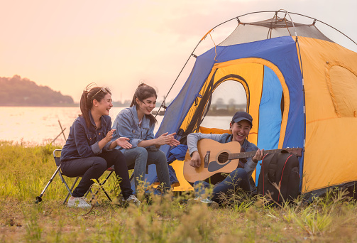 Group of Asian young woman friends having fun and enjoy in music of  guitar on parks and outdoor camping with lake background. Lifestyle and camping concept.