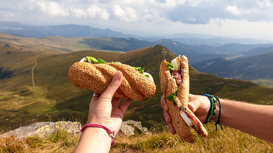 Lunch break in Austrian Alps. Two hands holding sandwiches with the view on high Alps. There are endless mountain chains in the back. Eating in the fresh air. A bit of overcast