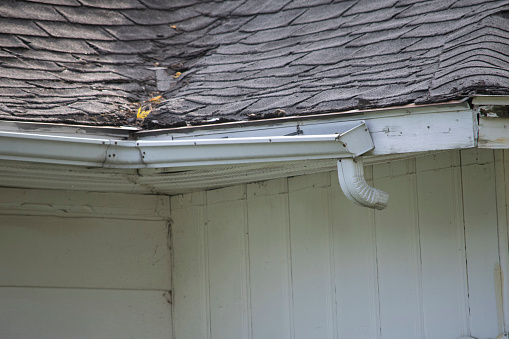 Roof of a residential house showing damage to the gutter systems, multiple layers of shingles, incomplete downspouts, and rusty gutters.