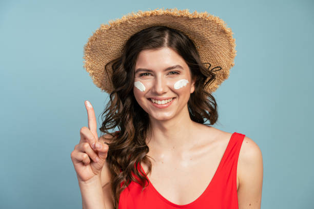 Beautiful, young woman in a straw hat applies sunscreen on her face. Cute girl posing on a blue background Beautiful, young woman in a straw hat applies sunscreen on her face. Cute girl posing on a blue background one piece swimsuit photos stock pictures, royalty-free photos & images