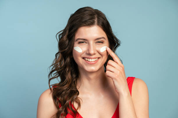 istock Smiling female wearing swimsuit and applying sunscreen lotion on face while looking at camera on blue background 1318702320