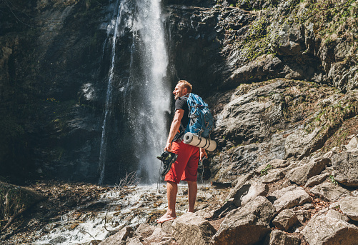 Hombre con mochila vestido con ropa activa de trekking sosteniendo botas de trekking en mano cerca de la cascada del río de montaña sonriendo y disfrutando de la naturaleza.Viajando, caminando, imagen conceptual de la naturaleza photo