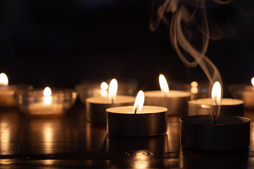 group of candles burning in the dark with light reflecting off of wood surface with one candle extinguished and smoke rising above it.  Copy space at top of image
