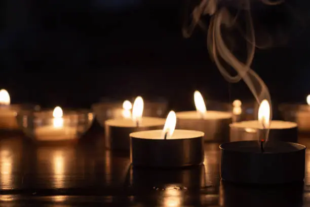 group of candles burning in the dark with light reflecting off of wood surface with one candle extinguished and smoke rising above it.  Copy space at top of image