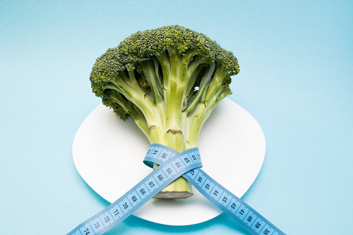 head of broccoli on a white plate wrapped in blue measuring tape, slimming and weight loss concept/ copy space top view