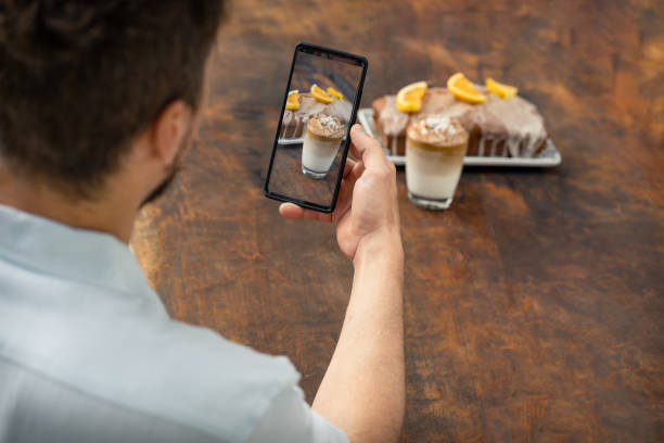 Man taking a photo with mobile at a glass of Dalgona coffee and an orange cake on a wooden background